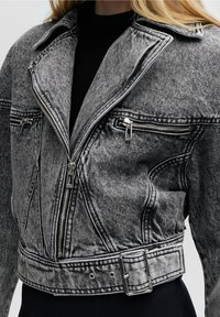 Grå denimmotorcykeljacka med en strukturerad design, dubbla blixtlås och bälte i midjan. Har en texturerad yta och framträdande s ömmar.