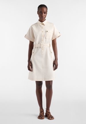 Kobieta ubrana w beżową sukienkę koszulową na krótki rękaw z paskiem, kieszeniami z przodu i guzikami, w połączeniu z brązowymi, rzymiankami na płaskiej podeszwie.