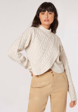 Maglione a trecce color crema con collo alto, caratterizzato da un design incrociato e maniche lunghe, indossato sopra pantaloni beige a vita alta.