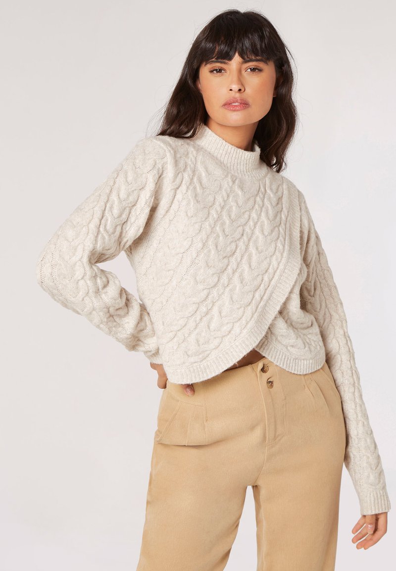 Creme farvet kabelstrikket sweater med høj hals, der har et krydset design og lange ærmer, båret over beige højtaljede bukser.