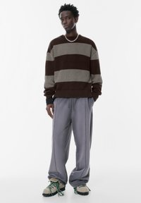 Pull en tricot rayé marron et gris associé à un pantalon large gris clair et des chaussures texturées avec une touche de couleur. Collier en chaîne argenté.