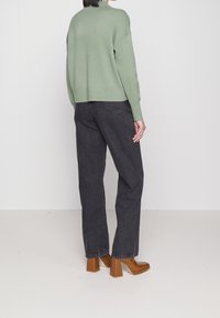 Grüner Strickpullover mit lockerer Passform, kombiniert mit hochgeschnittenen schwarzen Jeans und braunen Rundschaftstiefeln. Einfaches Design, minimale Details.