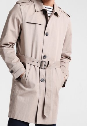 Trench-coat beige ceinturé avec des boutons, porté sur une chemise rayée par une personne les mains dans les poches, sur un fond clair.