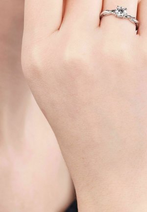 Hand met een zilveren verlovingsring met een ronde heldere diamant op de ringvinger tegen een bleke huidachtergrond.