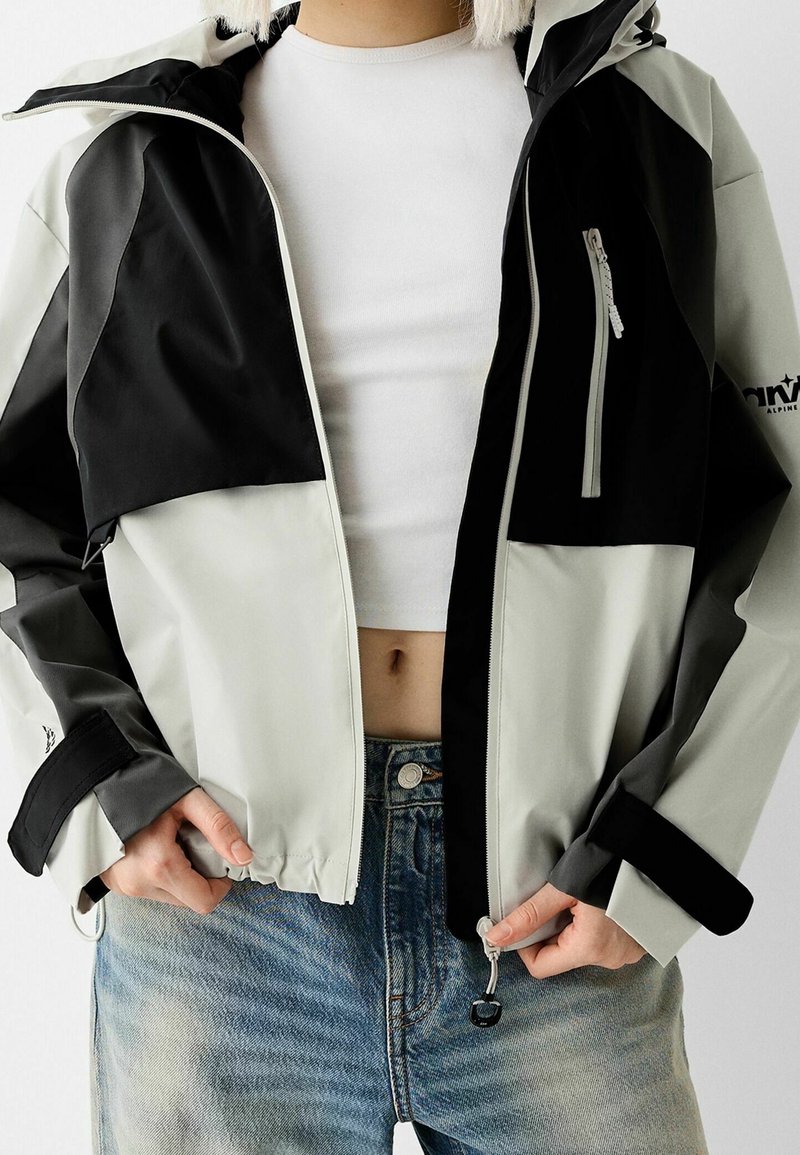 Personne portant une veste zippée noire et grise par-dessus un crop top blanc et un jean bleu, ajustant le fermoir éclair de la veste avec les deux mains.
