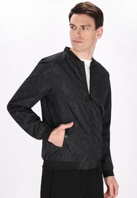 Giacca bomber nera con un motivo geometrico, texture liscia, polsini elastici e zip frontale. Presenta tasche laterali e un colletto rialzato.