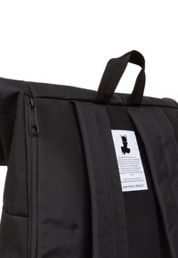 Svart tyg ryggsäck med ett handtag på toppen, dragkedjestängning och en etikett med ett grafiskt motiv. Slät textur och strukturerad design.