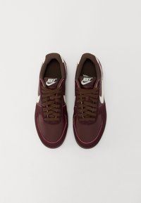 Vyno raudoni Nike sportbačiai su lygiu ir tekstūruotu dizainu, turintys rudus raištelius, baltą logotipą ir rožinius akcentus aplink padą.