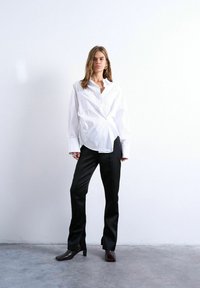Femme debout contre un mur blanc portant une chemise blanche ample boutonnée, un pantalon noir à jambes droites et des bottines à talons foncées.
