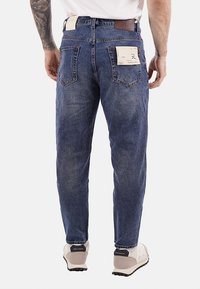 Jeans in denim blu con una vestibilità comoda, con una tasca in pelle marrone e due tasche posteriori. Cuciture visibili e finitura sbiadita.