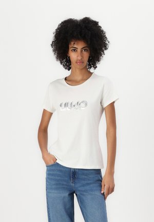 Cream cotton t-shirt with round neckline, short sleeves, and metallic logo detail on the front, paired with blue denim jeans.