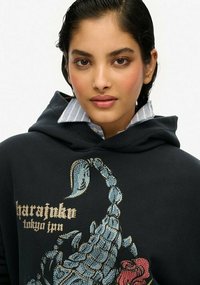 Felpa con cappuccio nera con una grafica di scorpione blu adornata con strass e dettagli floreali, indossata su una camicia a colletto blu chiaro.