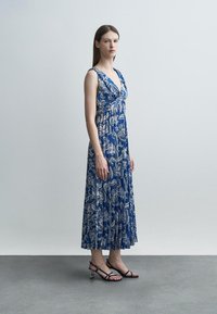 Žena stojaca v modro-bielych kvetovaných maxi šatách bez rukávov s plisovaním, obutá v čiernych sandáloch na podpätku so štýlovými remienkami, na neutrálnom pozadí.