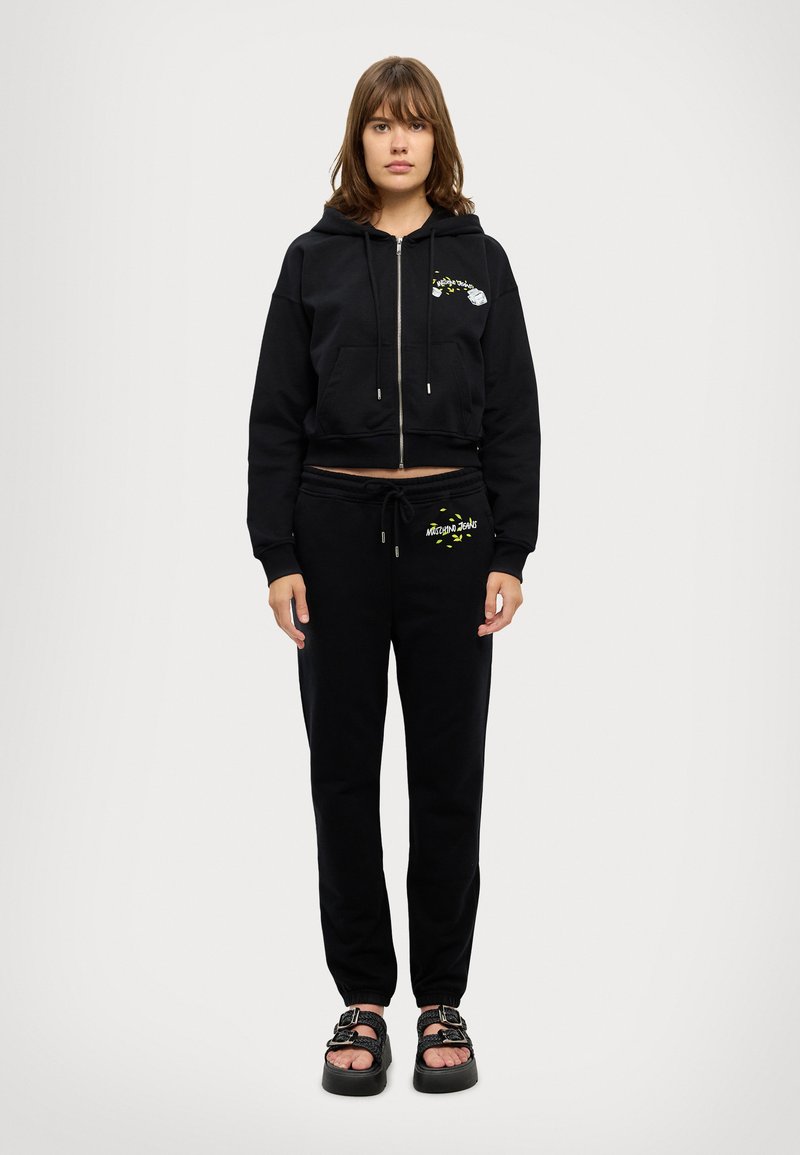 Schwarze cropped Hoodie mit Frontreißverschluss und Tasche, kombiniert mit schwarzen Jogginghosen mit Logo-Details. Das Model trägt schwarze Sandalen.