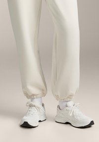 Białe sportowe sneakersy z teksturowanej siateczki i materiału skórzanego, z czarnymi akcentami, okrągłym noskiem i regulowanymi sznurowadłami. W zestawie z kremowymi zwężanymi joggerami.