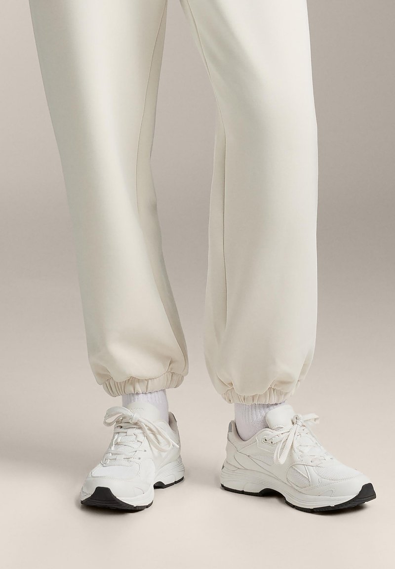 Białe sportowe sneakersy z teksturowanej siateczki i materiału skórzanego, z czarnymi akcentami, okrągłym noskiem i regulowanymi sznurowadłami. W zestawie z kremowymi zwężanymi joggerami.