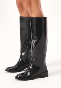 Bottes noires en cuir verni jusqu'au genou avec un bout arrondi, fermeture éclair sur le côté et un talon bloc pour la stabilité. Fini lisse et brillant.