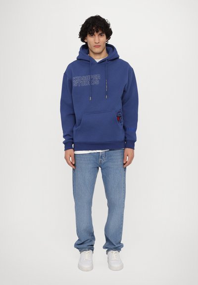 Felpa blu con tasca frontale, con la scritta "KIDSUPER STUDIOS" e una grafica di un leone rosso. Abbinata a jeans azzurri e sneakers bianche.
