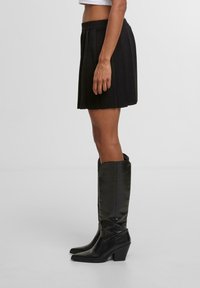 Černá skládací sukně s obepnutým pasem, spárovaná s černými koženými holinkami po kolena, které mají blokový podpatek a špičatou špičku.