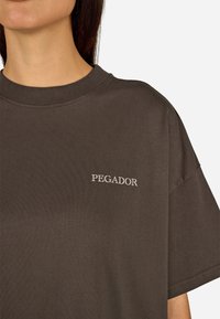 Brązowa bawełniana koszulka z okrągłym dekoltem i krótkimi rękawami; na lewej piersi wyszyte słowo "PEGADOR" na biało.