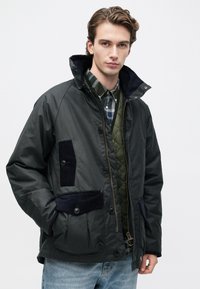 Tânăr îmbrăcat în geacă întunecată, cu straturi, vestă matlasată verde, cămașă în carouri și blugi deschiși, stând în fața unui fundal alb.