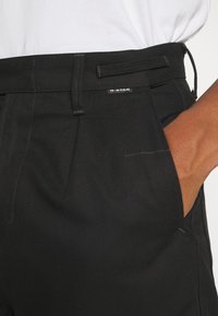 Close-up van een persoon die zwarte G-STAR broek draagt met een hand in de voorzak, gecombineerd met een wit ingestopt shirt.