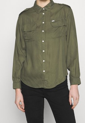 Osoba v olivově zelené košili na knoflíky se dvěma kapsami na hrudi a černých kalhotách, stojící před jednobarevným světlým pozadím.