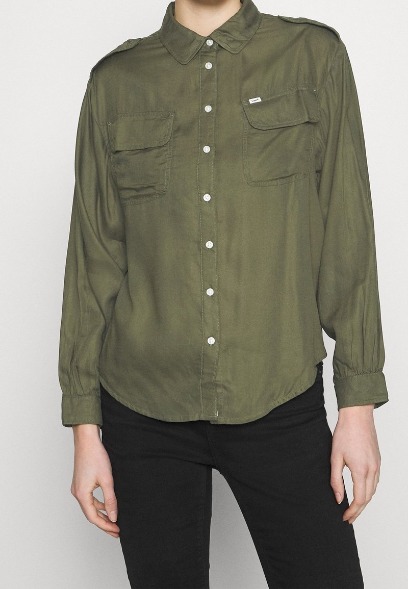 Person trägt ein olivgrünes Hemd mit Knöpfen und zwei Brusttaschen sowie schwarze Hose und steht vor einem schlichten hellen Hintergrund.