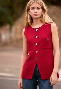 Top rosso senza maniche con taglio dritto, dotato di bottoni dorati e due tasche frontali. Realizzato in tessuto texturizzato, abbinato a jeans blu.