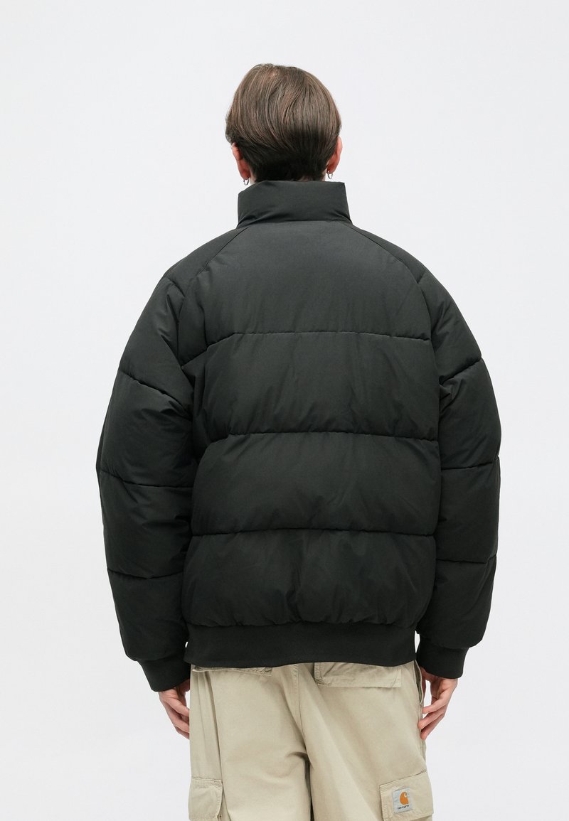 Giacca nera imbottita con colletto alto, caratterizzata da un design squadrato, sezioni cucite e polsini a coste. Indossata sopra pantaloni cargo beige.