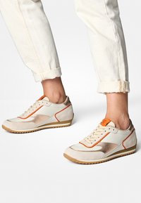 Baskets de sport crème et beige avec des accents orange, des détails métalliques, en daim texturé, semelle striée et design épuré.
