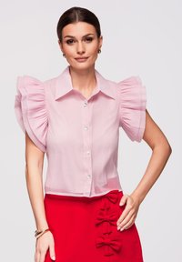 Femme portant un chemisier rose pâle avec des manches courtes volantées et une jupe rouge ornée de trois nœuds décoratifs.