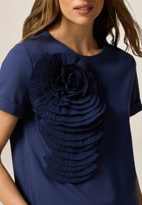 Marineblaue T-Shirt mit einem großen strukturierten, gefalteten Stoffdesign, das auf der linken Seite einer Rose ähnelt, rundem Ausschnitt und kurzen Ärmeln.