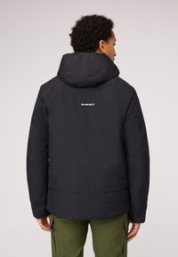 Giacca nera isolata con cappuccio, logo sulla schiena e texture liscia. Indossata con pantaloni cargo verdi.