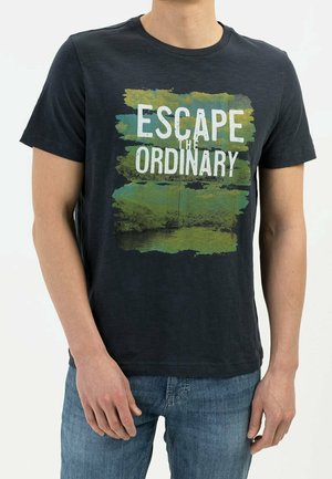 Man draagt een zwart T-shirt met witte tekst "ESCAPE THE ORDINARY" tegen een groene en blauwe abstracte achtergrond, gecombineerd met een blauwe spijkerbroek.