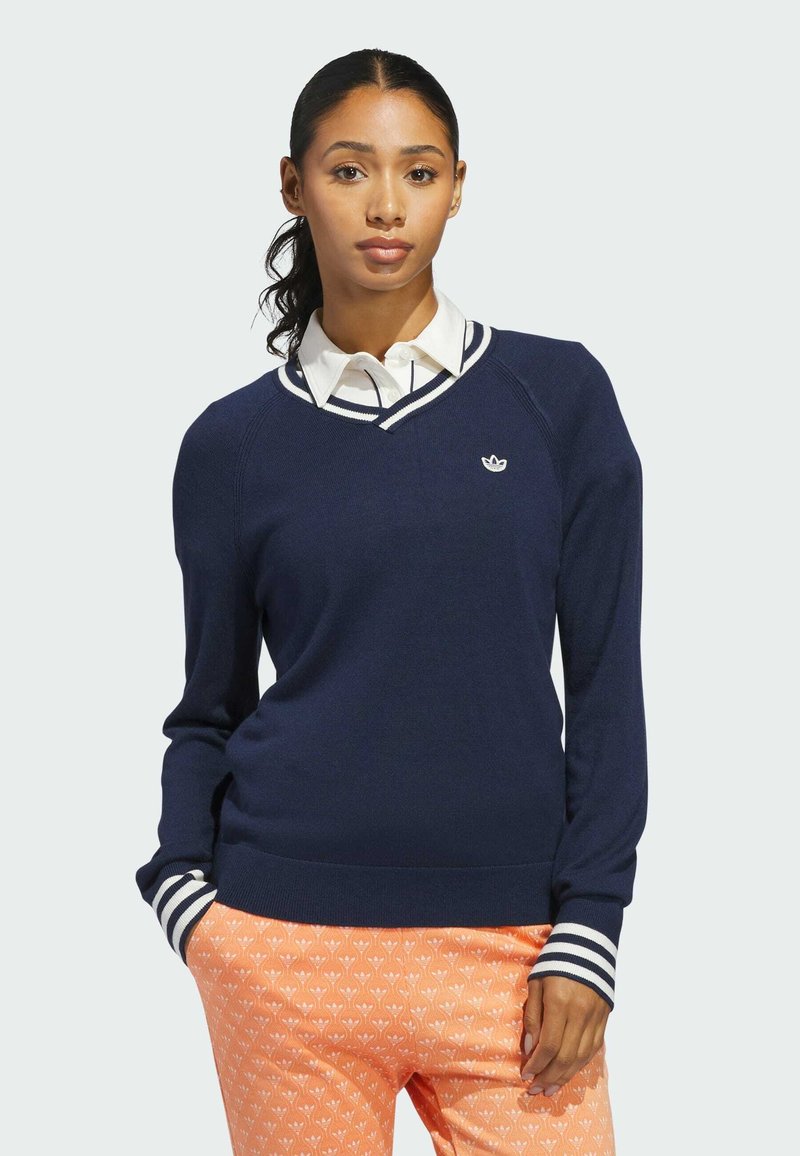 Μπλε πουλόβερ με V-λαιμόκοψη και λευκές ρίγες, με μικρό κεντημένο λογότυπο στο στήθος. Φορεμένο πάνω από πουκάμισο με γιακά.