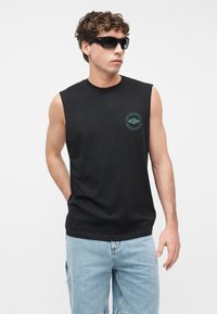 Νεαρός άνδρας που φοράει μαύρο αμάνικο μπλουζάκι με λογότυπο σερφ και ανοιχτό μπλε τζιν, με τα χέρια στις τσέπες, κοιτάζοντας πλάγια με γυαλιά ηλίου.