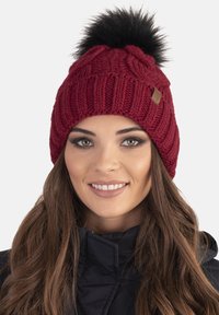 Gorro de punto burdeos con un gran pompom de piel sintética negra en la parte superior, que presenta una textura de canalé y un pequeño parche de logo marrón en el lateral.