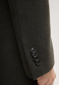 Donkergroene wollen jas met een gestructureerde afwerking, voorzien van vier zwarte knopen en subtiele stiksel-details langs de naad.