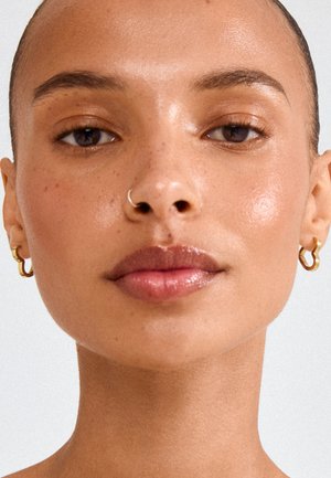 Close-up af en person med klar hud, guldringeøreringe, en næsering og subtil naturlig makeup mod en ensfarvet lys baggrund.