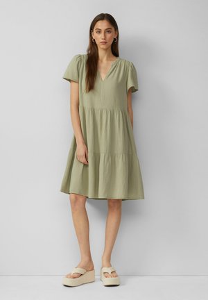 Hellgrünes Kleid mit V-Ausschnitt, kurzen Puffärmeln, gestuftem Rock und strukturiertem Stoff. Dazu werden beige Plateausandalen getragen.
