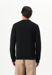 Homem com cabelo escuro a usar um suéter preto de mangas longas e calças bege, visto de costas contra um fundo branco liso.