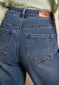 Sötétkék farmernadrág magas derékkal, két hátsó zsebbel és barna bőrből készült márkajelzéssel a derékszár felett. Texturált anyag.