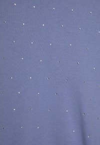 Tessuto azzurro chiaro con borchie di metallo argentato sparse, creando un motivo testurizzato su una superficie liscia. Il materiale appare morbido ed elastico.