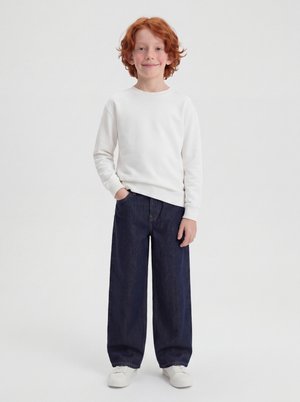 Rødhåret barn stående iført en hvid langærmet skjorte, brede mørkeblå bukser, hvide sneakers, med den ene hånd i lommen.