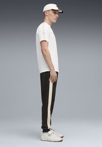 Hvid t-shirt med mønstret skulderkanter, sorte bukser med guldstriber i siden, og hvide sneakers med lysebrune accenter.