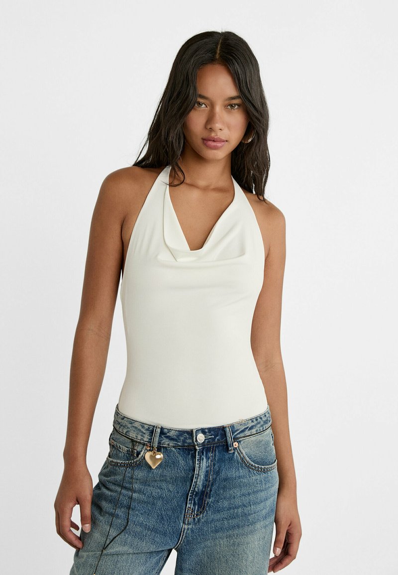Body blanc sans manches avec un col drapé, associé à un jean en denim baggy bleu clair avec un charm en forme de cœur sur la boucle de ceinture.