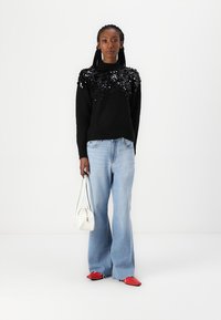 Maglione nero con dettagli in paillettes sulle spalle, abbinato a jeans a gamba larga blu chiaro. Tiene in mano una piccola borsa bianca e indossa scarpe rosse.