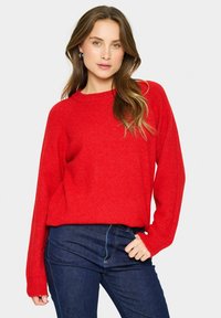 Roter Strickpullover mit rundem Ausschnitt, langen Ärmeln und gerippten Bündchen, kombiniert mit dunkelblauen Jeans. Einfache Textur, keine Muster oder Verzierungen.