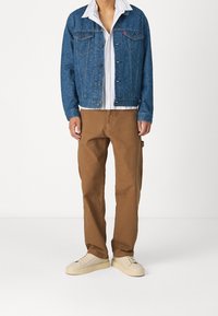 Jeansjacka i blått med ett mönstrat textur, vit skjorta under, och bruna cargobyxor med en avslappnad passform. Ljusfärgade sneakers.